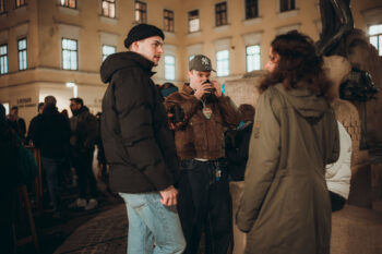 Drei Personen stehen nachts auf einem Platz in der Stadt, der mit anderen Menschen gefüllt ist, und unterhalten sich. Ein Mann trägt eine schwarze Jacke und eine Mütze, ein anderer trinkt aus einer Tasse, und eine Frau trägt einen grünen Mantel. Im Hintergrund sind Gebäude mit beleuchteten Fenstern zu sehen.