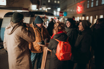 Eine Gruppe warm gekleideter Menschen steht nachts um einen hohen Tisch im Freien, plaudert und lächelt auf einer Stadtstraße mit festlichen Lichtern und Imbissbuden im Hintergrund.