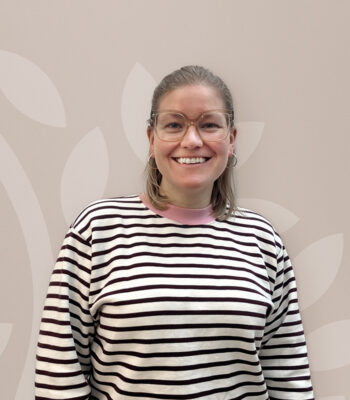 Eine Person mit schulterlangem blondem Haar, Brille und Ohrringen lächelt in die Kamera. Sie trägt einen schwarz-weiß gestreiften Pullover mit rosa Kragen von VABON und steht vor einem hellen, gemusterten Hintergrund.