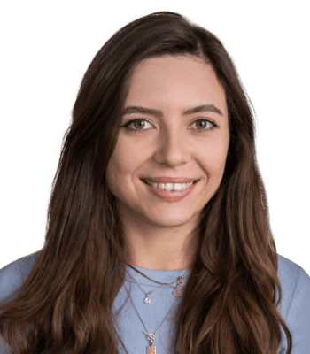 Eine junge Social Media Managerin mit langen braunen Haaren lächelt in die Kamera. Sie trägt ein hellblaues Oberteil und mehrlagige Ketten und steht vor einem schlichten weißen Hintergrund.