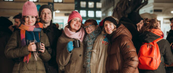 Fünf Personen in warmer Winterkleidung lächeln und posieren zusammen bei einer Abendveranstaltung im Freien, jede hält einen Becher SPiNNpunsch. Im Hintergrund sind andere Menschen und festliche Lichter zu sehen.