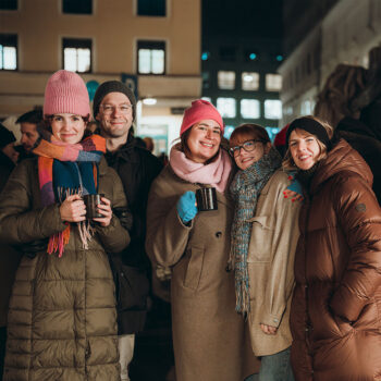 Fünf Personen in warmen Wintermänteln und Mützen lächeln und posieren nachts zusammen und halten Becher mit SPiNNpunsch bei einer Veranstaltung im Freien. Im Hintergrund sind städtische Gebäude mit beleuchteten Fenstern zu sehen.