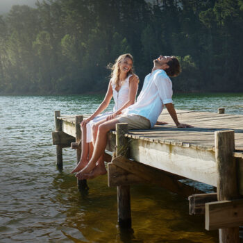 Ein junges Paar sitzt auf einem hölzernen Steg an einem See, lächelt und lacht zusammen, während ihre Füße über dem Wasser baumeln. Im Hintergrund sind hohe Bäume und Sonnenlicht zu sehen, die eine friedliche Szene schaffen, über die der sanfte Geist von Guardi wacht.