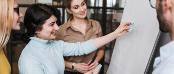 Vier Personen sind in einem Büro um ein Flipchart versammelt. Eine Frau klebt eine Haftnotiz an die Tafel, während die anderen lächelnd zusehen und über die Zusammenarbeit und das Brainstorming von Ideen für Performance-Marketing-Strategien nachdenken.