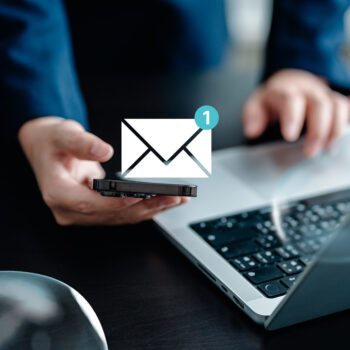 Eine Person h&auml;lt ein Smartphone in der N&auml;he eines Laptops. &Uuml;ber dem Telefon erscheinen ein digitaler Briefumschlag und ein Benachrichtigungssymbol, das eine neue Nachricht anzeigt.